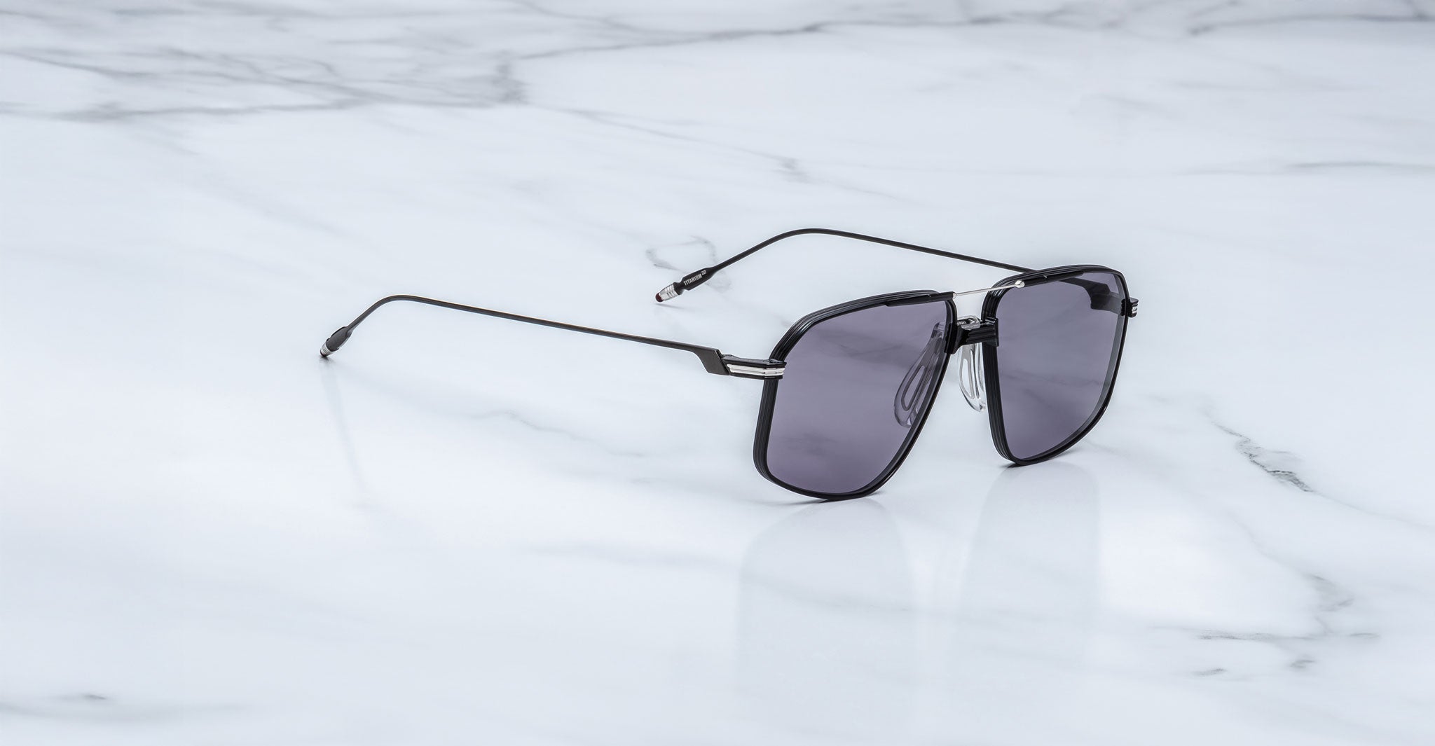A pair of Jacques Marie Mage Jagger sunglasses by Jacques Marie Mage, featuring an oversized aviator shape with dark lenses and beta titanium accents, rests on a white marble surface.