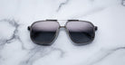 The Jacques Marie Mage Jagger sunglasses by Jacques Marie Mage, featuring black square beta titanium metal frames, rest on a white marble surface.