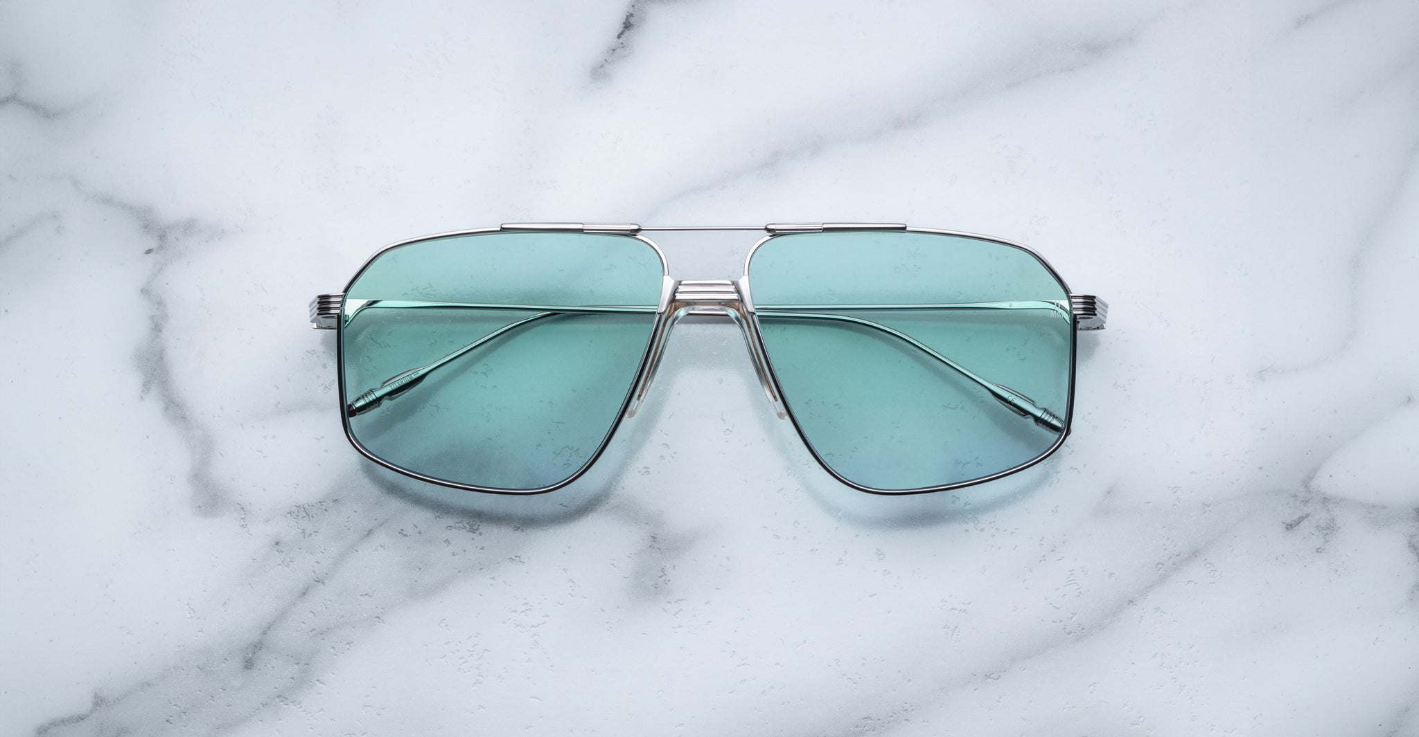 A pair of Jacques Marie Mage Jagger oversized aviator sunglasses with teal-tinted lenses and metal frames from Jacques Marie Mage rests on a white marble surface.