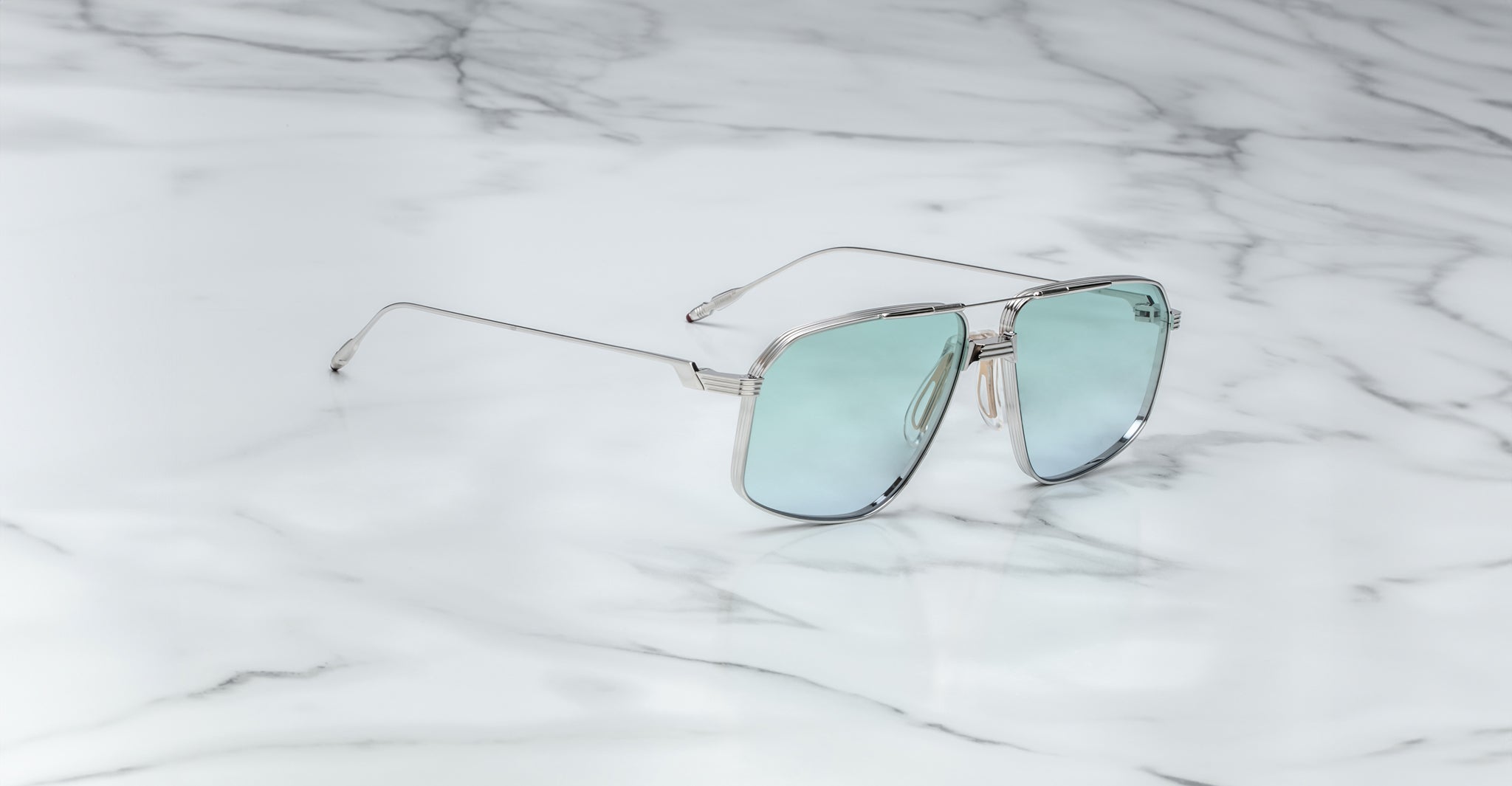 The Jacques Marie Mage Jagger sunglasses from Jacques Marie Mage feature an oversized aviator design with a silver metal frame, light blue reflective lenses, and beta titanium construction, shown elegantly on a white marble surface.