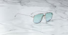 The Jacques Marie Mage Jagger sunglasses from Jacques Marie Mage feature an oversized aviator design with a silver metal frame, light blue reflective lenses, and beta titanium construction, shown elegantly on a white marble surface.