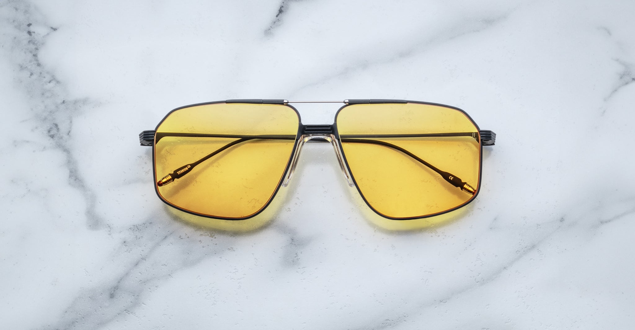 A pair of Jacques Marie Mage Jagger oversized aviator sunglasses with yellow-tinted lenses and sleek black arms rests on a white marble surface.