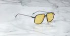 A pair of Jacques Marie Mage Jagger oversized aviator sunglasses with black frames and yellow lenses by Jacques Marie Mage sits on a white marble surface.