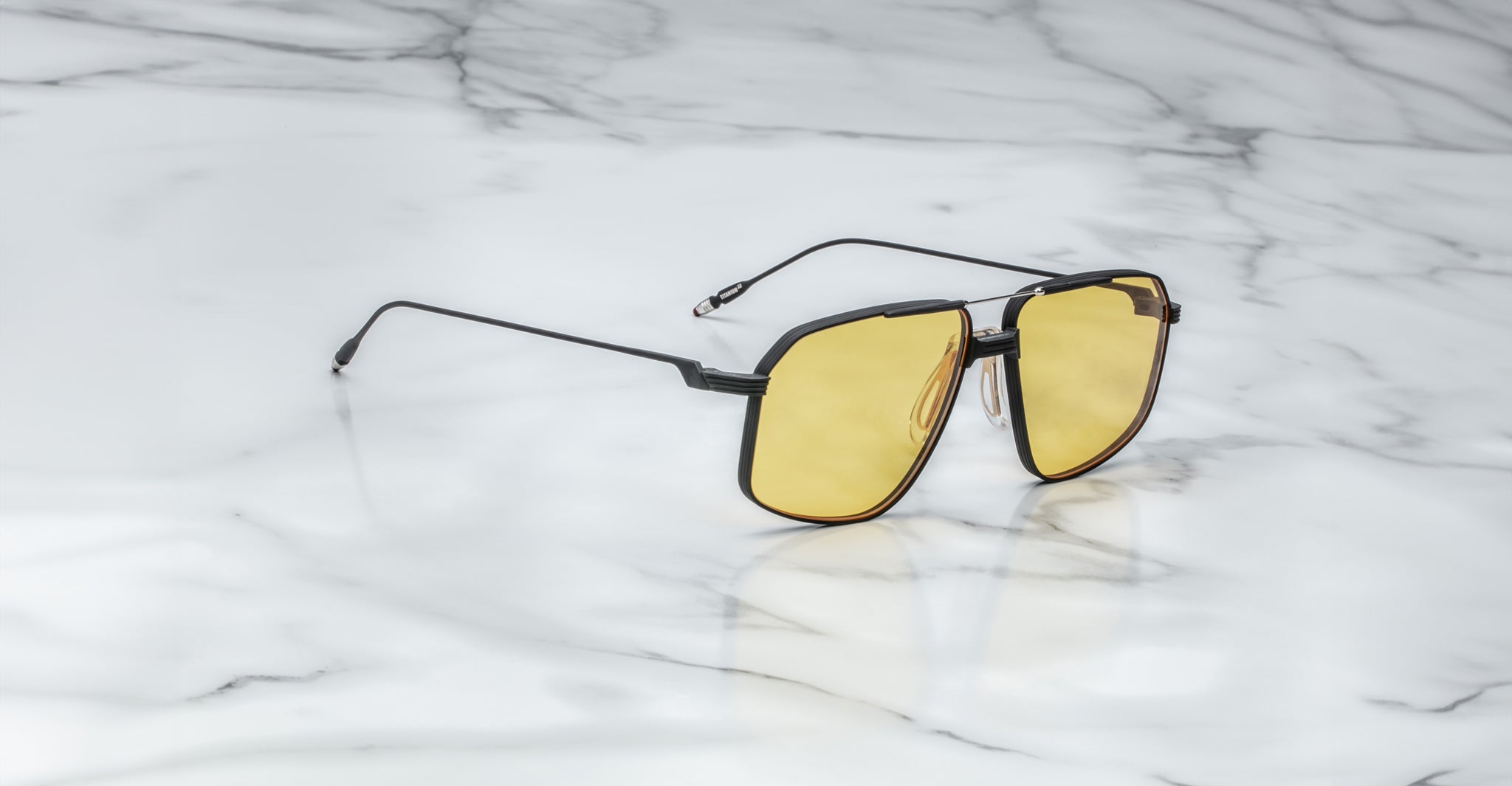 A pair of Jacques Marie Mage Jagger oversized aviator sunglasses with black frames and yellow lenses by Jacques Marie Mage sits on a white marble surface.