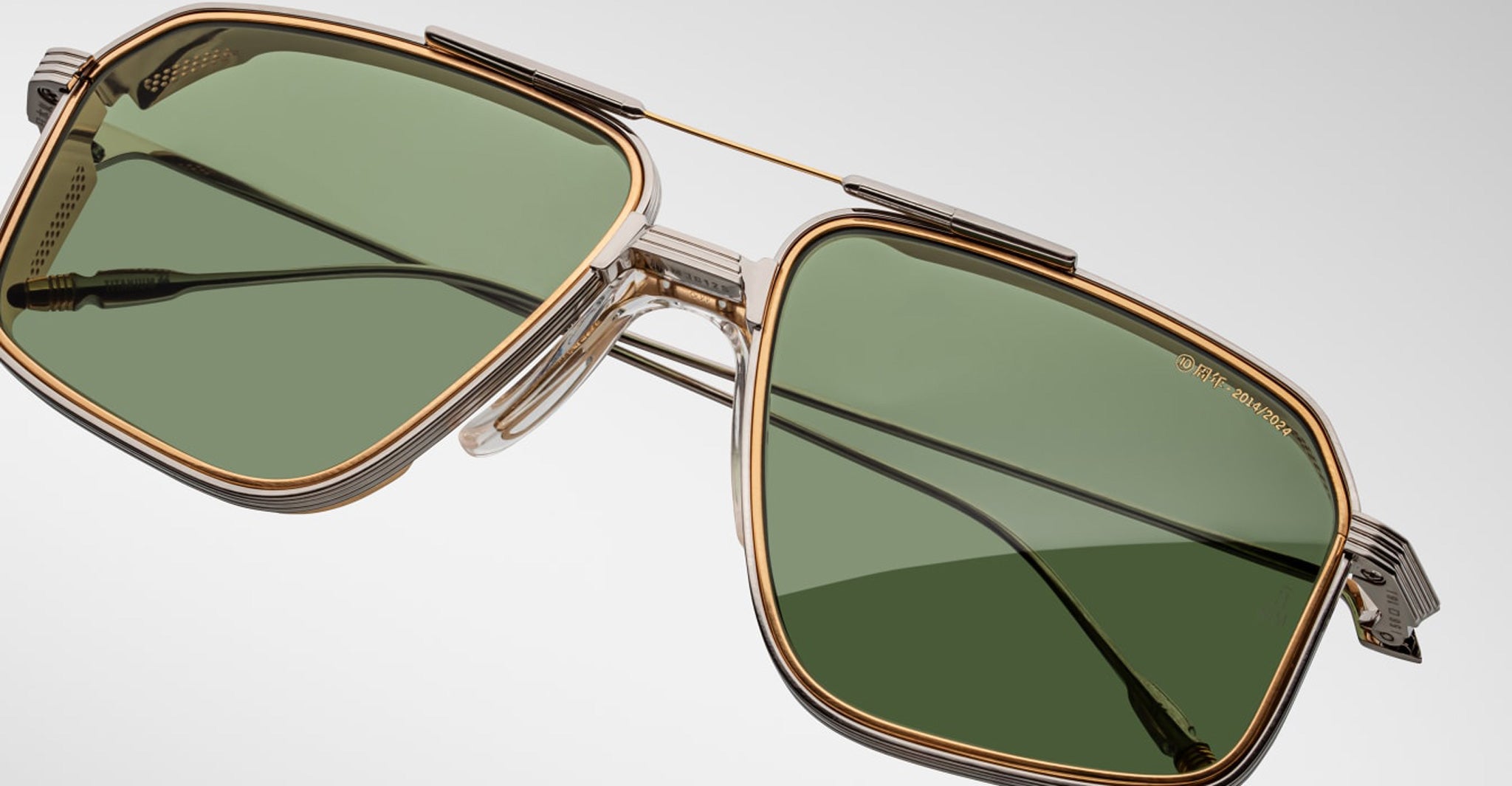 Close-up of Jacques Marie Mage Jagger sunglasses by Jacques Marie Mage, featuring green-tinted rectangular lenses, gold and silver beta titanium frames, and black earpieces against a plain background.