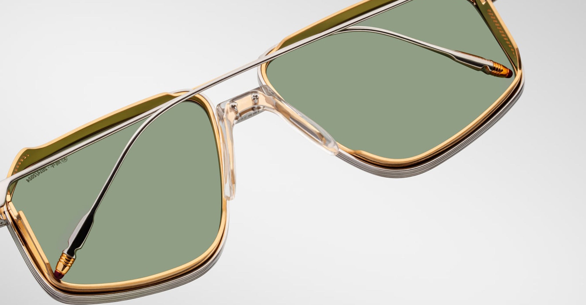 Close-up of Jacques Marie Mage Jagger aviator sunglasses by Jacques Marie Mage, featuring oversized transparent and gold frames, green-tinted lenses, and silver beta titanium arms, displayed on a light background.