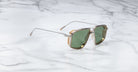 The Jacques Marie Mage Jagger sunglasses by Jacques Marie Mage feature oversized aviator frames with green lenses, gold accents, and a beta titanium metal frame, displayed stylishly on a white marble surface.