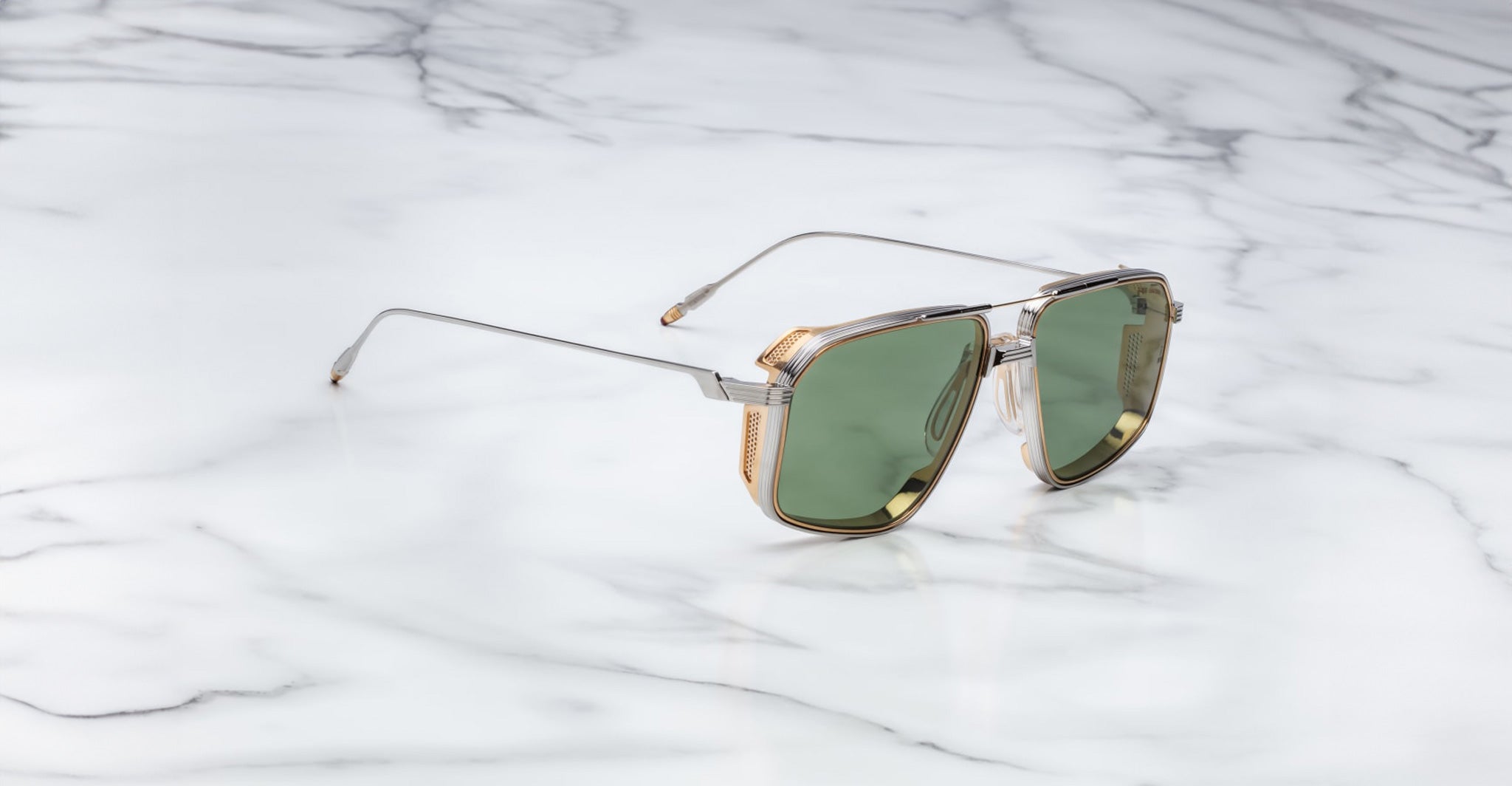 The Jacques Marie Mage Jagger sunglasses by Jacques Marie Mage feature oversized aviator frames with green lenses, gold accents, and a beta titanium metal frame, displayed stylishly on a white marble surface.