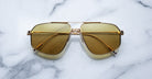The Jagger sunglasses by Jacques Marie Mage, with oversized gold beta titanium frames and yellow-tinted lenses, are displayed on a white marble surface.