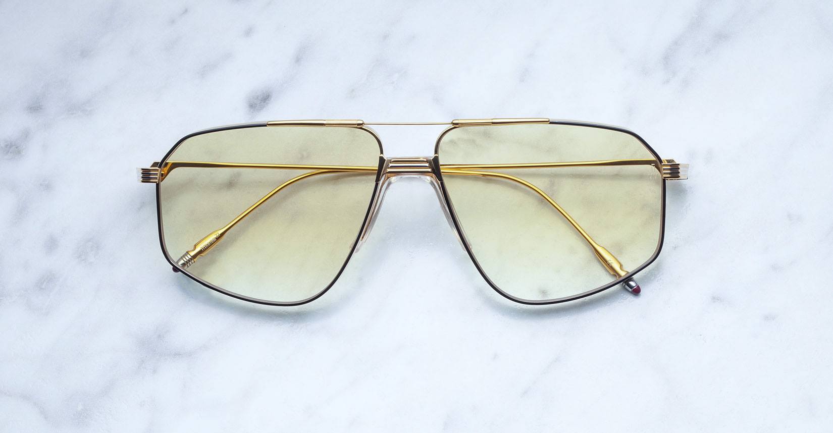 The Jacques Marie Mage Jagger sunglasses, featuring oversized gold aviator frames and yellow-tinted lenses, rest elegantly on a white marble surface.