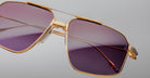 Close-up of Jacques Marie Mage Jagger oversized aviator sunglasses by Jacques Marie Mage, featuring gold frames and purple gradient lenses, displayed against a plain gray background.