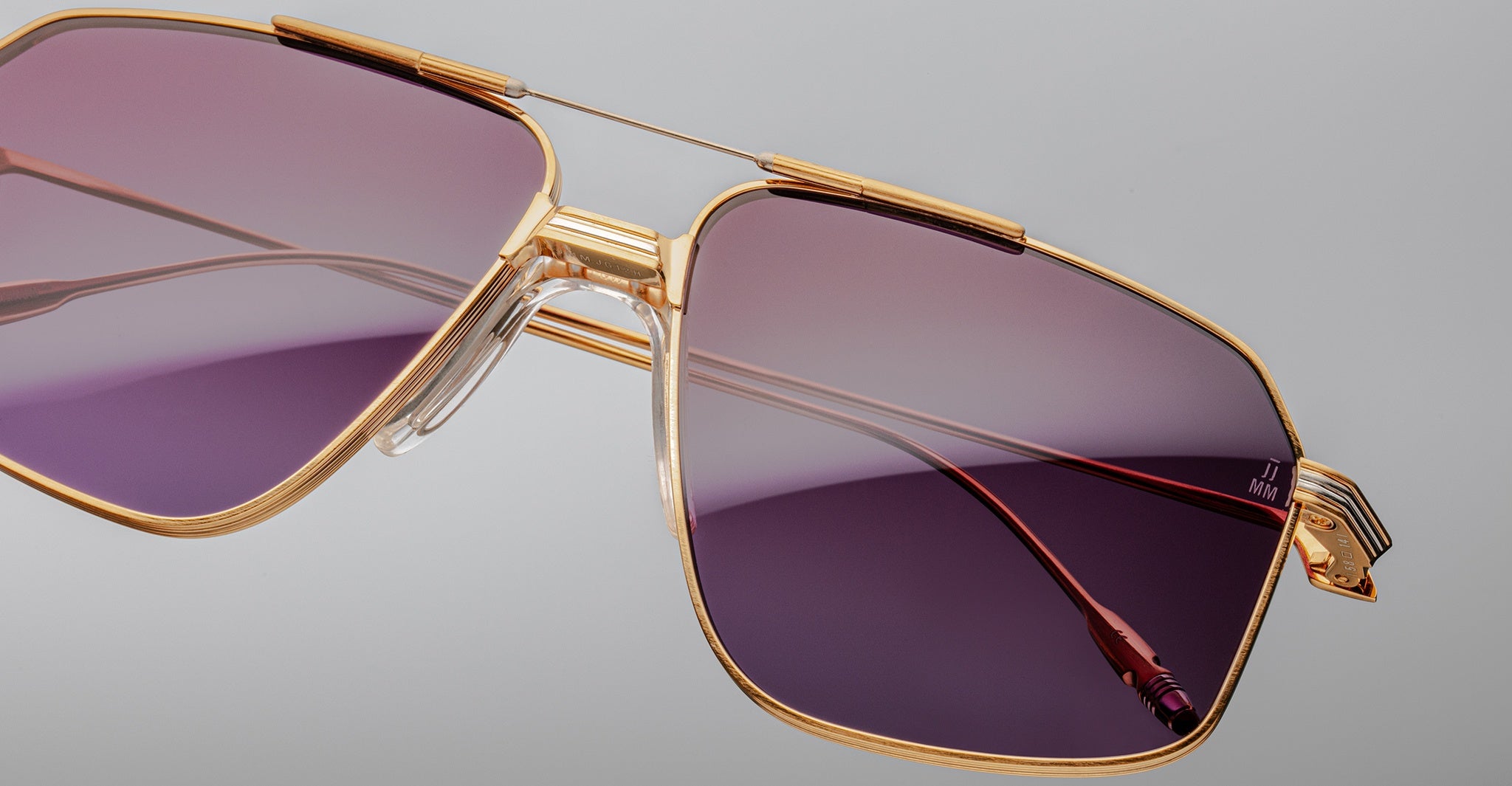 Close-up of Jacques Marie Mage Jagger oversized aviator sunglasses by Jacques Marie Mage, featuring gold frames and purple gradient lenses, displayed against a plain gray background.