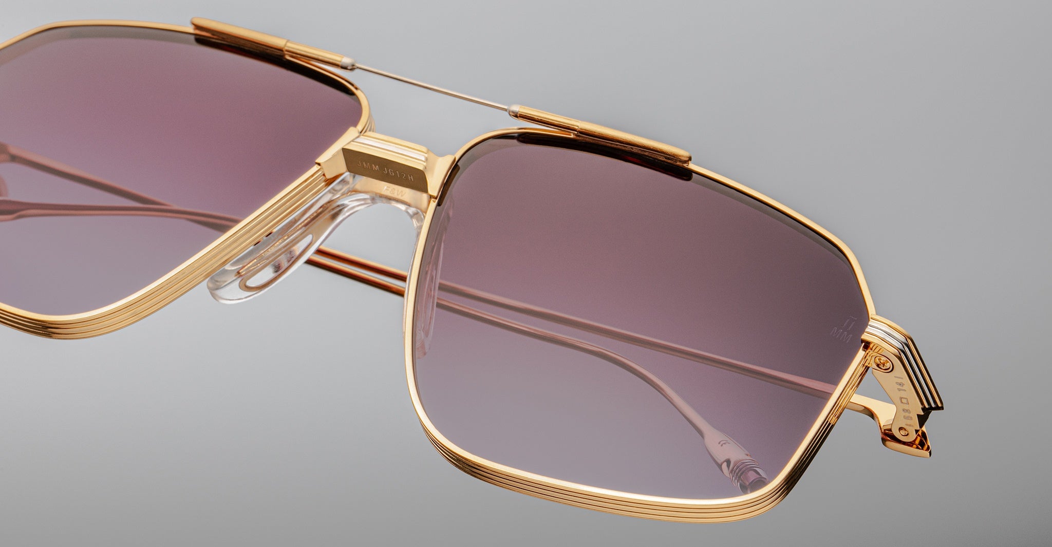 Close-up of oversized Jacques Marie Mage Jagger aviator sunglasses by Jacques Marie Mage, featuring gold beta titanium frames, purple-tinted lenses, clear nose pads, and detailed hinges, set against a plain gray background.
