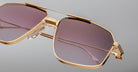 Close-up of oversized Jacques Marie Mage Jagger aviator sunglasses by Jacques Marie Mage, featuring gold beta titanium frames, purple-tinted lenses, clear nose pads, and detailed hinges, set against a plain gray background.