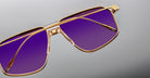 Close-up of Jacques Marie Mage Jagger oversized aviator sunglasses by Jacques Marie Mage, with gold beta titanium frames and purple-tinted lenses, shown on a gray background.