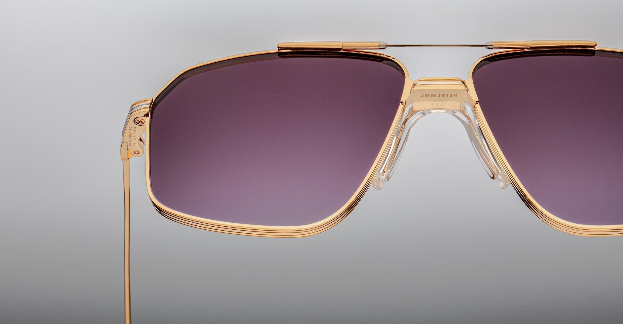 Close-up of Jacques Marie Mage Jagger oversized aviator sunglasses by Jacques Marie Mage, featuring gold frames and purple-tinted lenses, set against a plain light gray background.