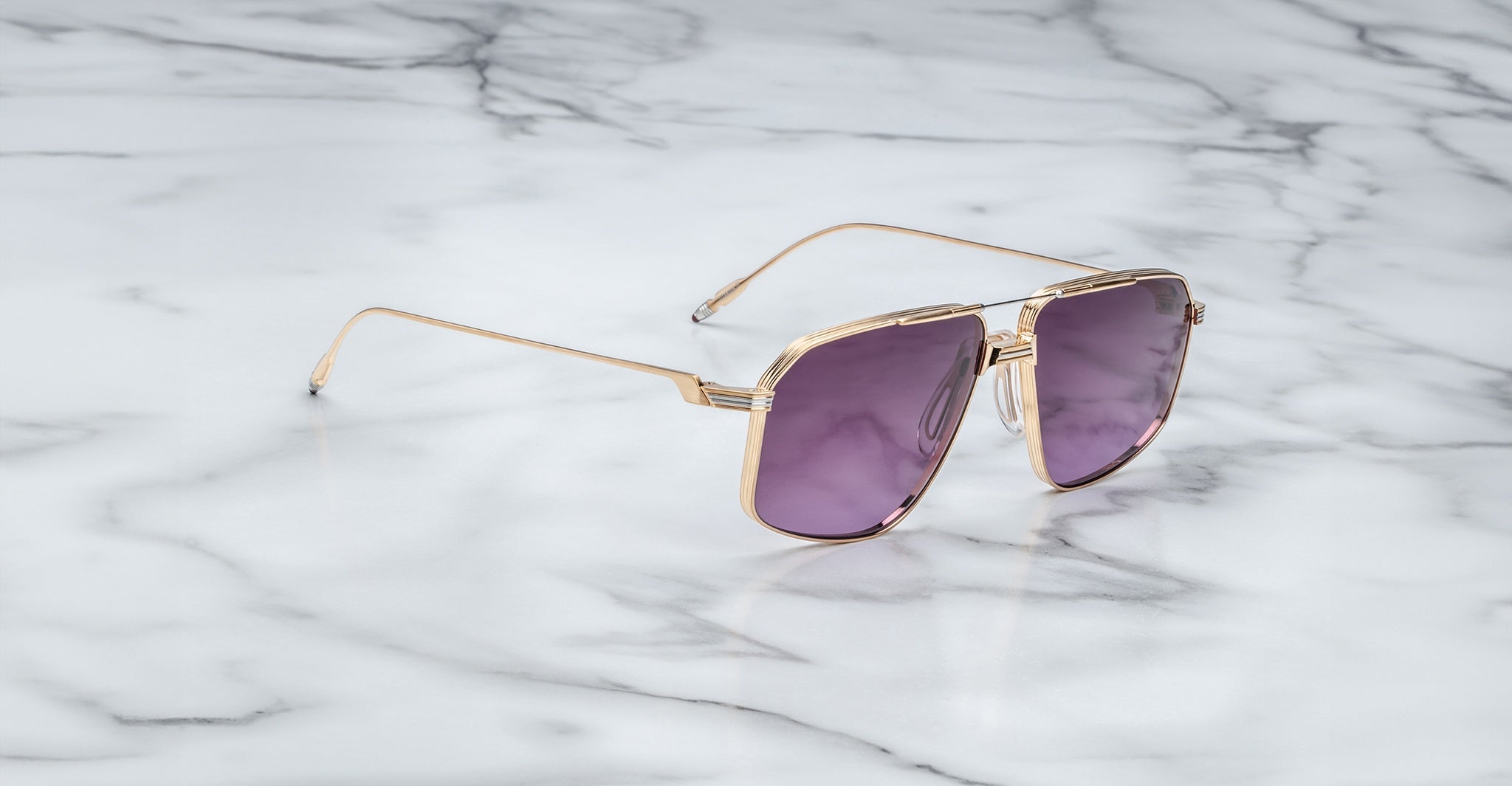 The Jacques Marie Mage Jagger sunglasses by Jacques Marie Mage, featuring gold frames and purple lenses, are displayed elegantly on a white marble surface.
