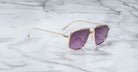 The Jacques Marie Mage Jagger sunglasses by Jacques Marie Mage, featuring gold frames and purple lenses, are displayed elegantly on a white marble surface.