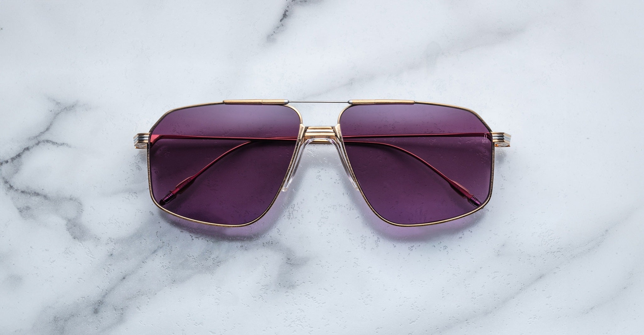 A pair of Jacques Marie Mage Jagger sunglasses with gold frames and purple lenses by Jacques Marie Mage sits on a white marble surface.