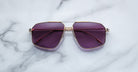 A pair of Jacques Marie Mage Jagger sunglasses with gold frames and purple lenses by Jacques Marie Mage sits on a white marble surface.