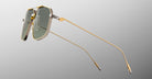 The Jacques Marie Mage Jagger by Jacques Marie Mage features gold-framed, rectangular sunglasses inspired by oversized aviators, crafted from beta titanium with green-tinted lenses, shown on a grey gradient background.