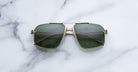 A pair of Jacques Marie Mage Jagger gold-framed, green-tinted oversized aviator sunglasses from Jacques Marie Mage is displayed on a white marble surface.