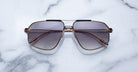 The Jacques Marie Mage Jagger sunglasses feature oversized aviator-style brown gradient lenses and slim beta titanium frames by Jacques Marie Mage, displayed on a white marble surface.