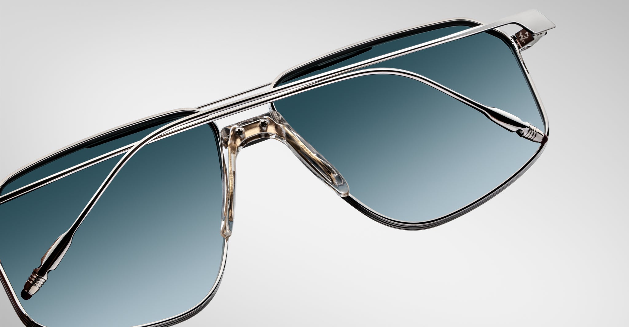 Close-up of Jacques Marie Mage Jagger aviator sunglasses by Jacques Marie Mage, featuring oversized sleek beta titanium frames and blue gradient lenses on a light gray background.