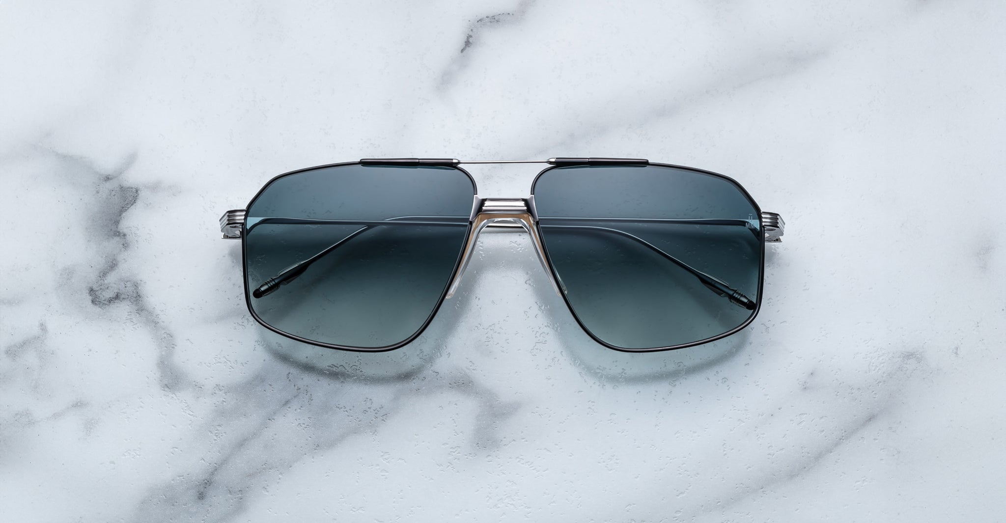The Jacques Marie Mage Jagger by Jacques Marie Mage features oversized aviator sunglasses with dark tinted lenses and a sleek beta titanium frame, elegantly displayed on a white marble surface.