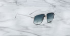 Jacques Marie Mage Jagger aviator sunglasses with silver frames and blue gradient lenses by Jacques Marie Mage rest on a white marble surface.