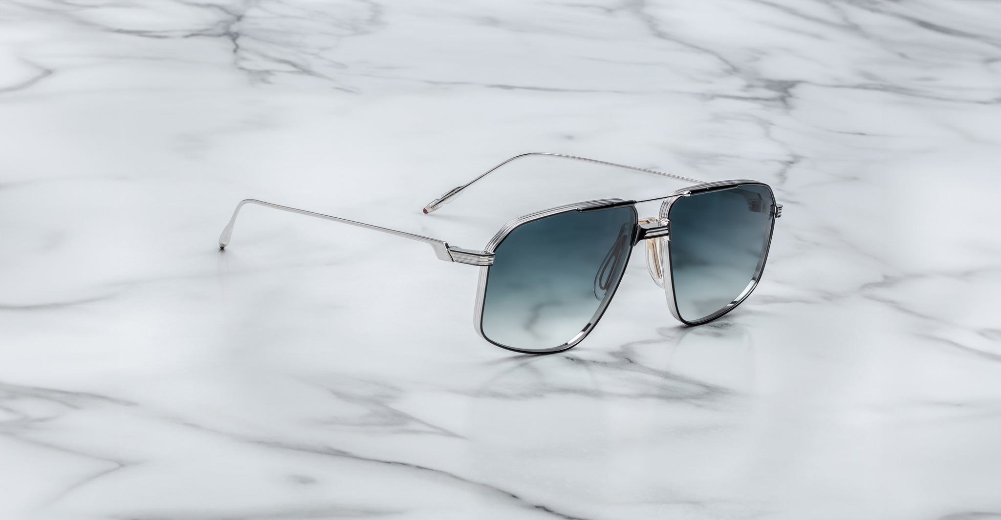 Jacques Marie Mage Jagger aviator sunglasses with silver frames and blue gradient lenses by Jacques Marie Mage rest on a white marble surface.