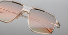 Close-up of the Jacques Marie Mage Jagger sunglasses by Jacques Marie Mage, featuring oversized gold beta titanium frames and gradient pink-tinted lenses, shown against a plain gray background.