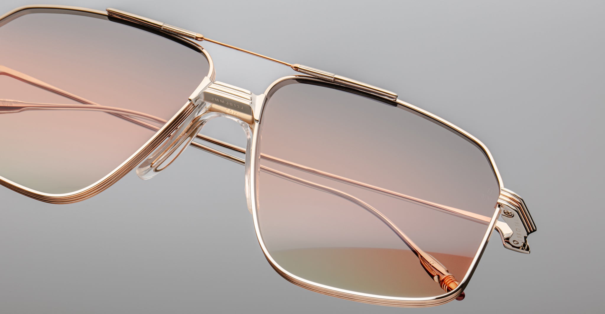 Close-up of the Jacques Marie Mage Jagger sunglasses by Jacques Marie Mage, featuring oversized gold beta titanium frames and gradient pink-tinted lenses, shown against a plain gray background.