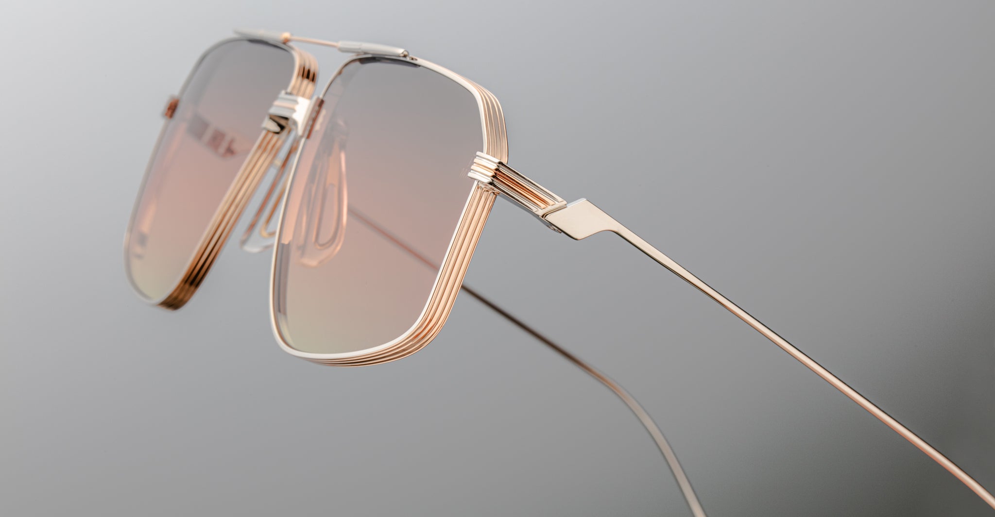 Close-up of Jacques Marie Mage Jagger sunglasses by Jacques Marie Mage, showcasing oversized aviator style with gold metal frames, pink-tinted lenses, and beta titanium construction against a neutral gray background.