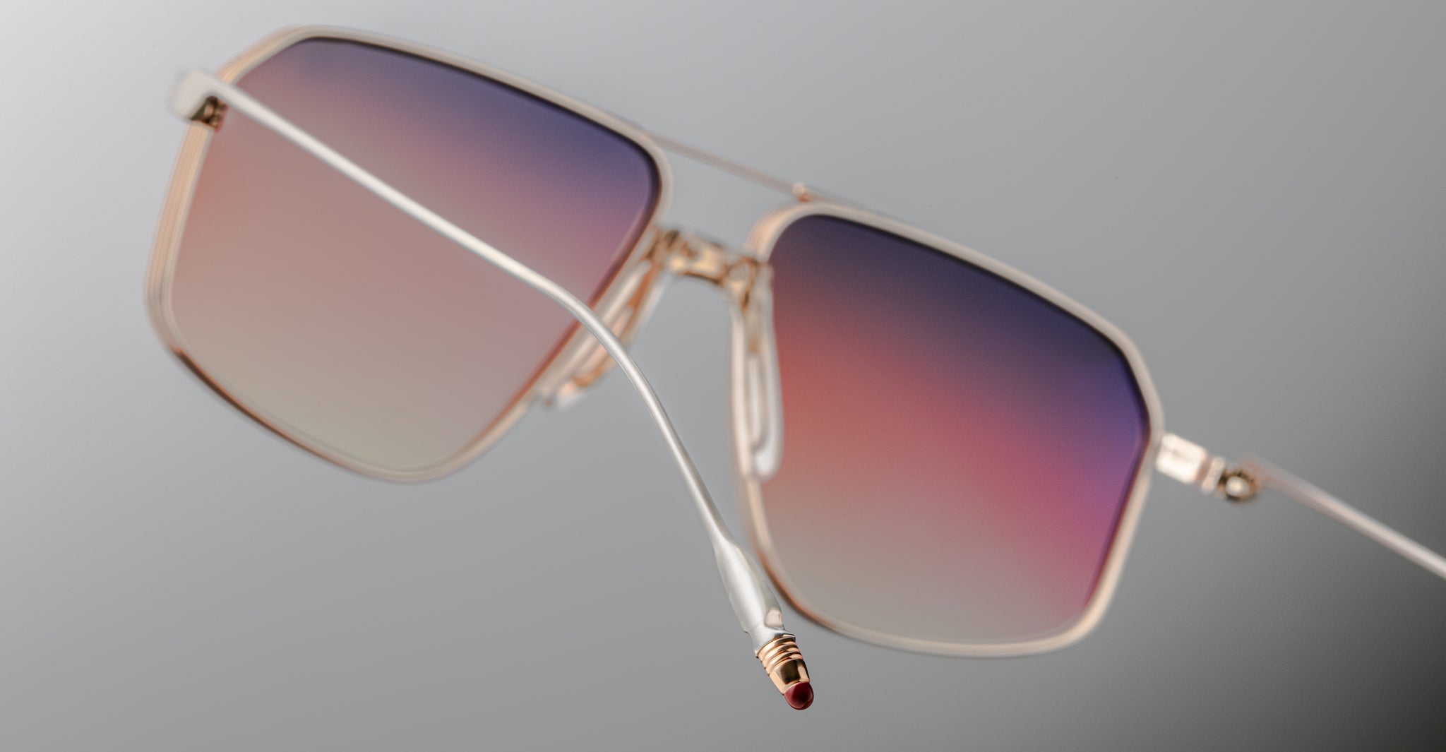 Close-up of Jacques Marie Mage Jagger sunglasses by Jacques Marie Mage, featuring gold rectangular beta titanium frames and gradient lenses fading from purple to pink, set against a neutral background.