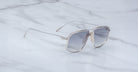 The Jacques Marie Mage Jagger silver metal oversized aviator sunglasses with gray-tinted lenses rest elegantly on a white marble surface.