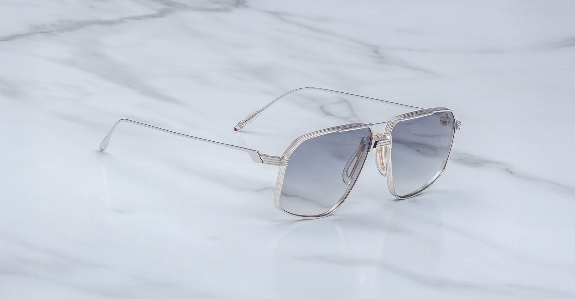 The Jacques Marie Mage Jagger silver metal oversized aviator sunglasses with gray-tinted lenses rest elegantly on a white marble surface.
