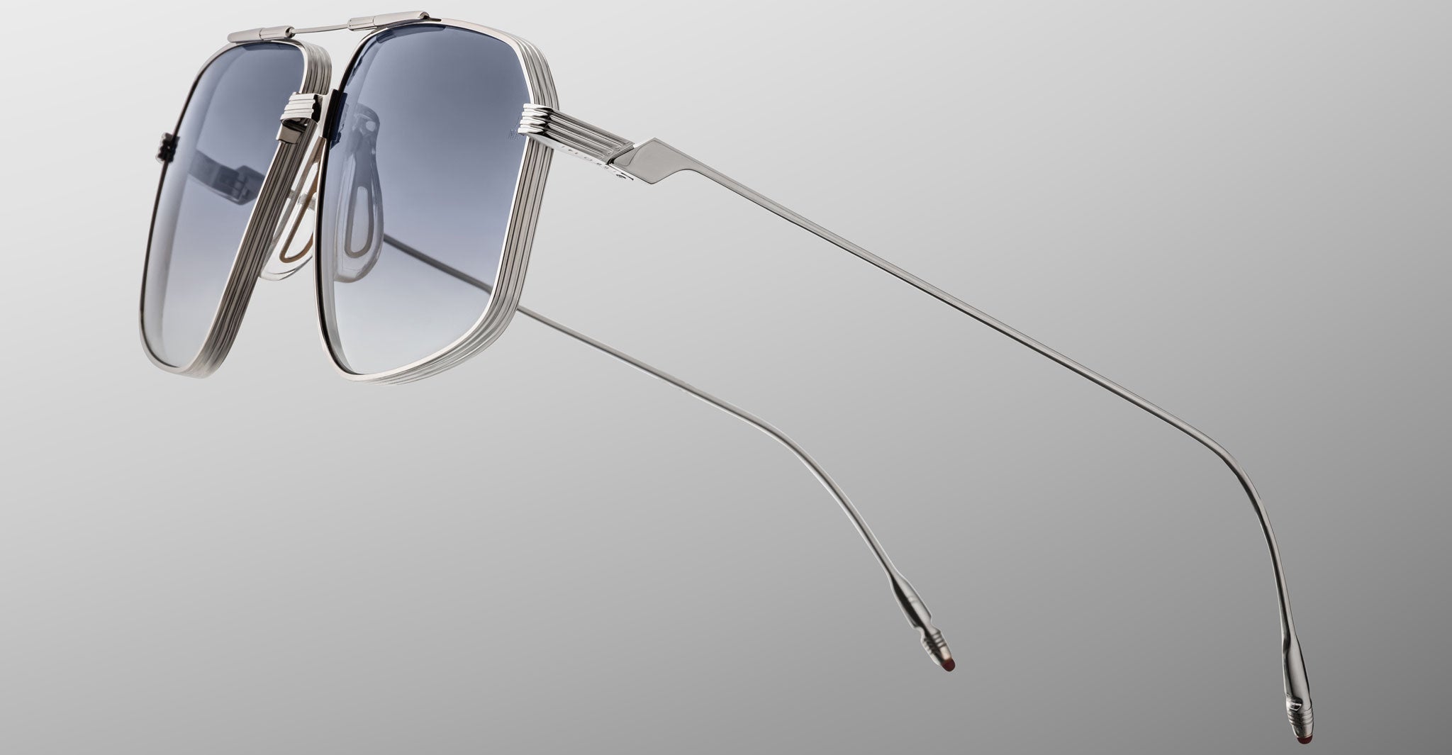 The Jacques Marie Mage Jagger sunglasses by Jacques Marie Mage feature silver oversized aviator frames with blue-tinted lenses, shown against a gray gradient background and angled to showcase thin beta titanium arms.