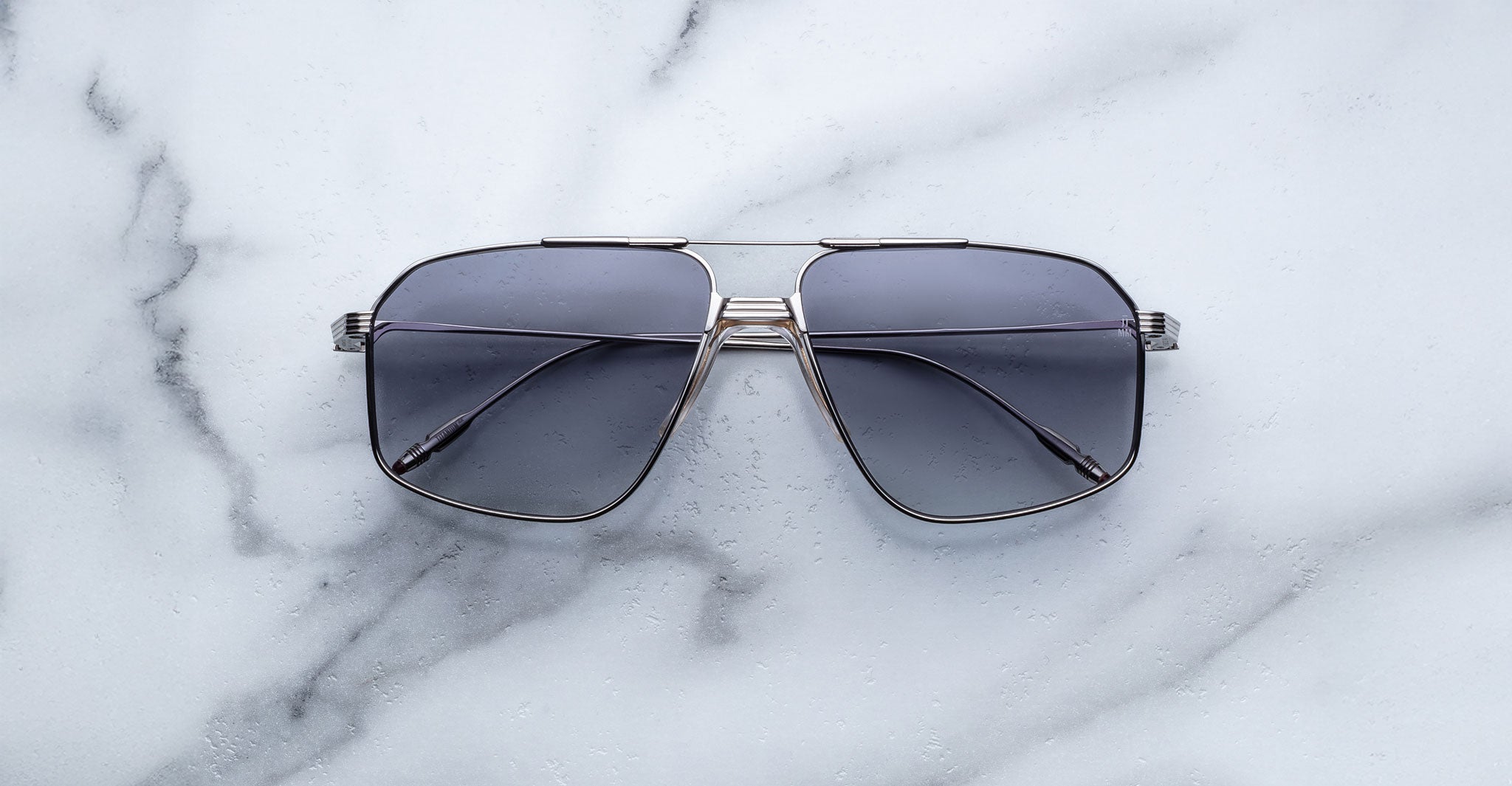 A pair of Jacques Marie Mage Jagger oversized aviator sunglasses with dark lenses and sleek metal frames from Jacques Marie Mage rests on a light marble surface.
