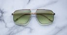 The Jacques Marie Mage Jagger sunglasses by Jacques Marie Mage, featuring gold frames and green-tinted oversized aviator lenses, rest elegantly on a white marble surface.