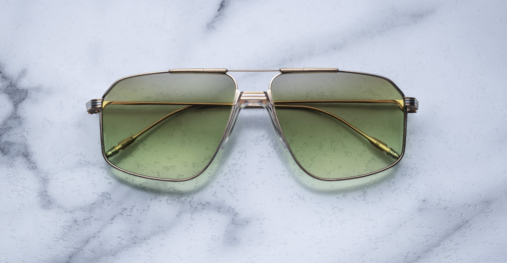 The Jacques Marie Mage Jagger sunglasses by Jacques Marie Mage, featuring gold frames and green-tinted oversized aviator lenses, rest elegantly on a white marble surface.