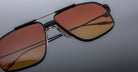 Close-up of Jacques Marie Mage Jagger sunglasses: oversized aviator style with black frames, gradient orange-brown lenses, and beta titanium accents on the bridge, nose pads, and temples, set against a grey background.