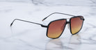 The Jacques Marie Mage Jagger sunglasses feature square black frames and gradient orange lenses, inspired by the signature style of Jacques Marie Mage, and are elegantly displayed on a white marble surface.