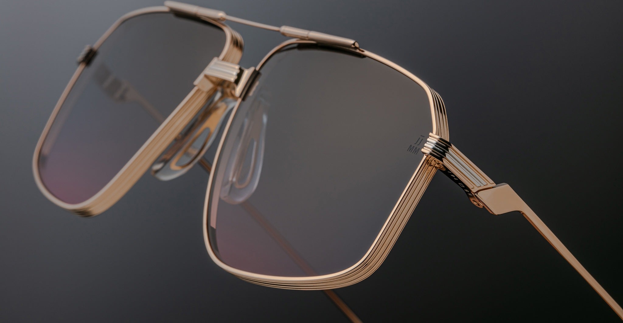 Close-up of Jacques Marie Mage Jagger aviator sunglasses by Jacques Marie Mage, featuring a gold geometric beta titanium frame, tinted lenses, and prominent nose pads, inspired by the iconic Jagger style against a dark background.