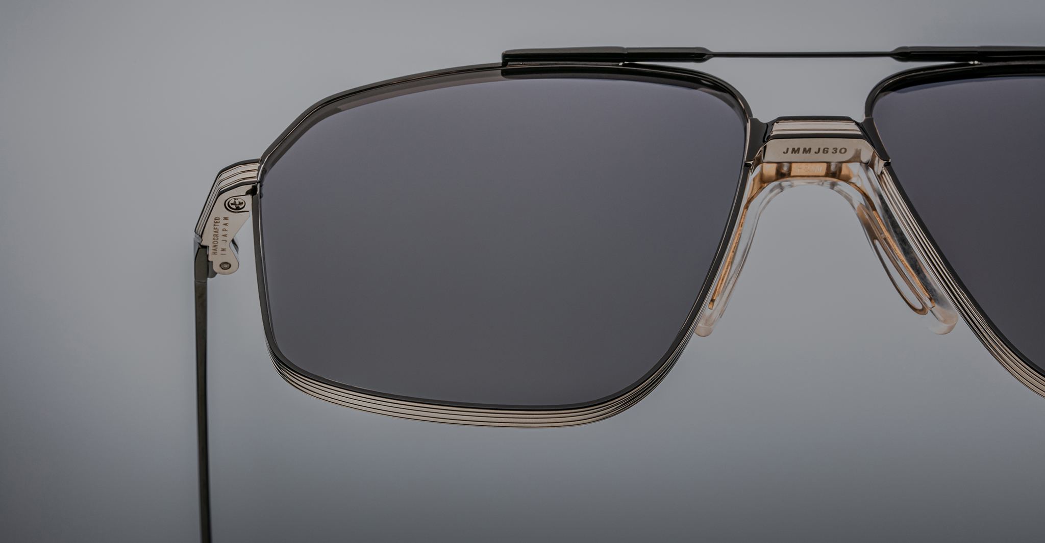 Close-up of Jacques Marie Mage Jagger sunglasses by Jacques Marie Mage, featuring oversized aviator style, dark lenses, a beta titanium metal frame, and visible hinge detail on a neutral gray background.