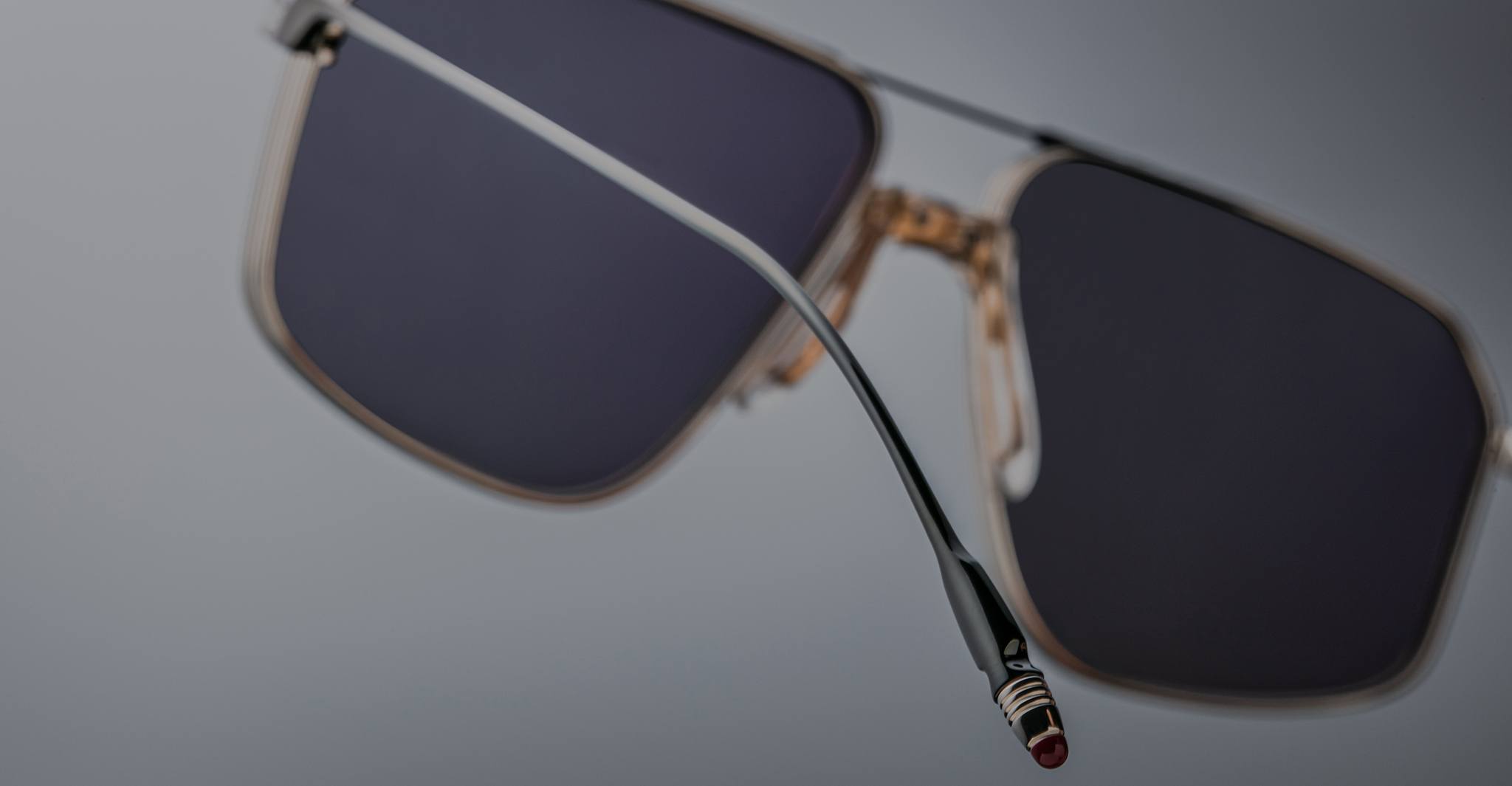 Close-up of Jacques Marie Mage Jagger sunglasses by Jacques Marie Mage, featuring black rectangular frames, gold accents, beta titanium details, and a red temple tip against a blurred gray background.