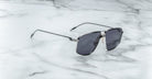 A pair of Jacques Marie Mage Jagger oversized aviator sunglasses with dark lenses and beta titanium accents rests stylishly on a white marble surface, reflecting the bold elegance of the Jacques Marie Mage brand.