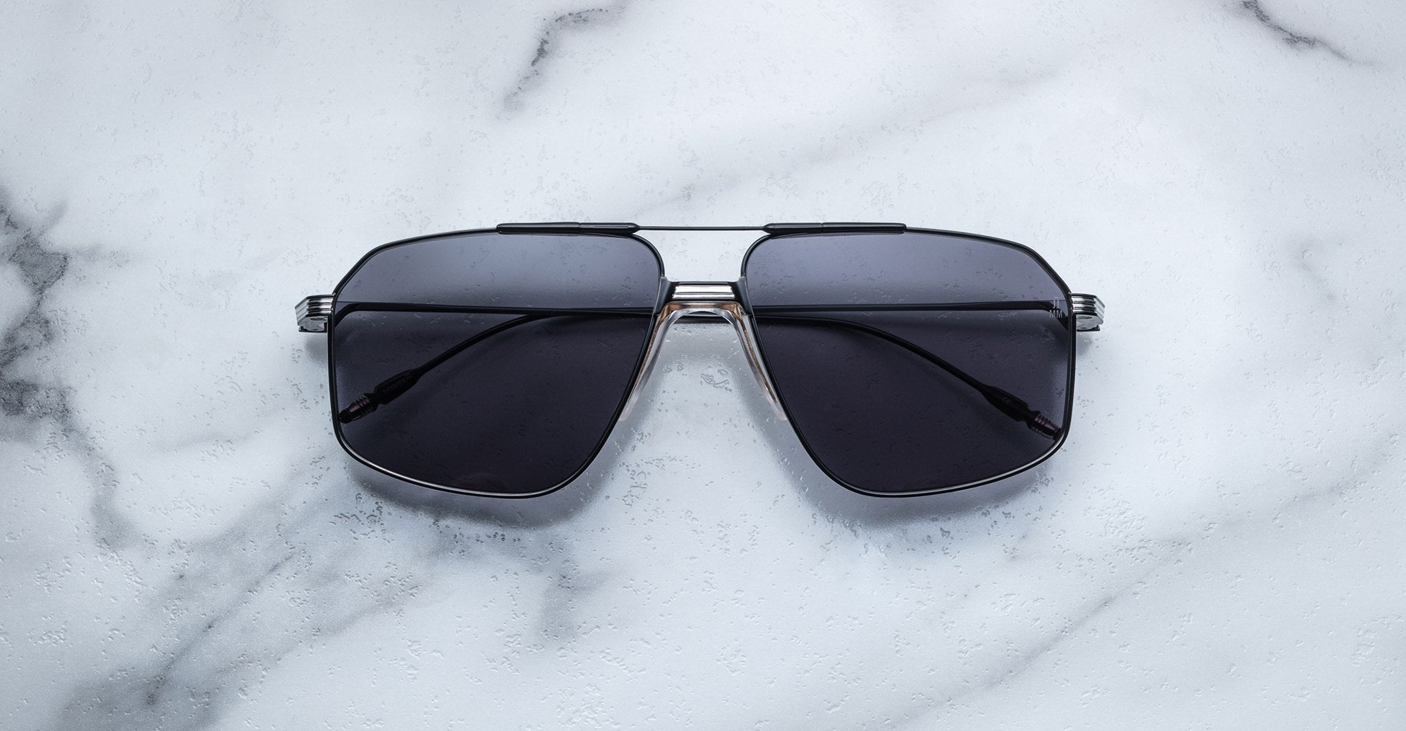 Jacques Marie Mage Jagger oversized aviator sunglasses by Jacques Marie Mage, featuring dark lenses and a silver beta titanium frame, are displayed on a white marble surface.
