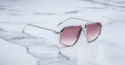 A pair of Jacques Marie Mage Jagger oversized aviator sunglasses by Jacques Marie Mage, featuring thin beta titanium frames and pink-tinted lenses, sits on a white marble surface.