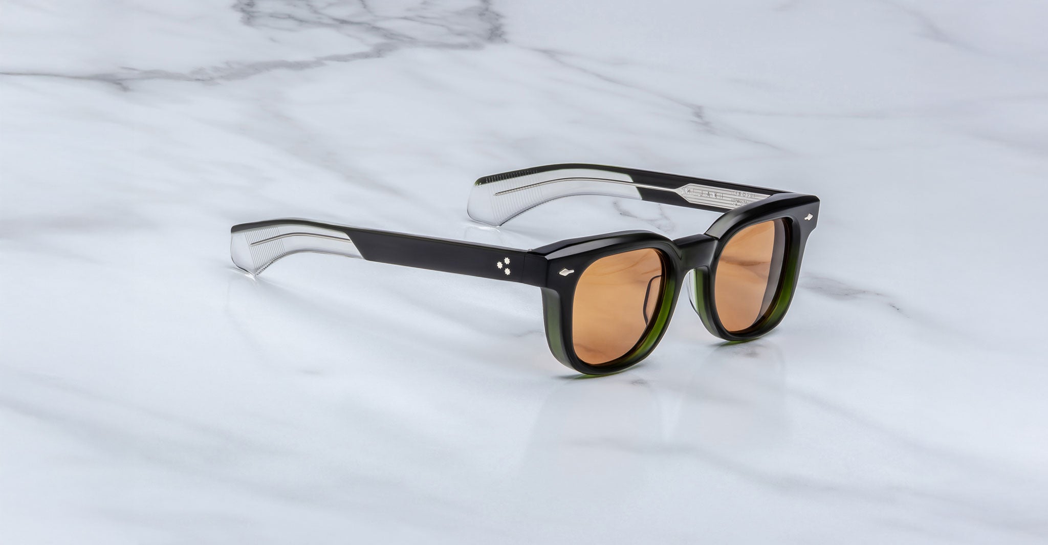 A pair of Jacques Marie Mage Jax 2 sunglasses, featuring black and clear acetate with brown-tinted lenses and gradient-effect arms, rests on a white marble surface.